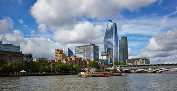 This urban landscape photograph shows River Boomerang floating on the River Thames in London during the morning in summer. The scene is characterised by distinctive architecture along the riverfront, including modern buildings and residential blocks. The prominent glass-clad skyscraper One Blackfriars, also known as the Boomerang, rises above the surrounding structures and serves as a well-known London landmark in the image. The bright summer morning light highlights the details of the riverside architecture and the bridge crossing the Thames, emphasising the urban nature of the setting.